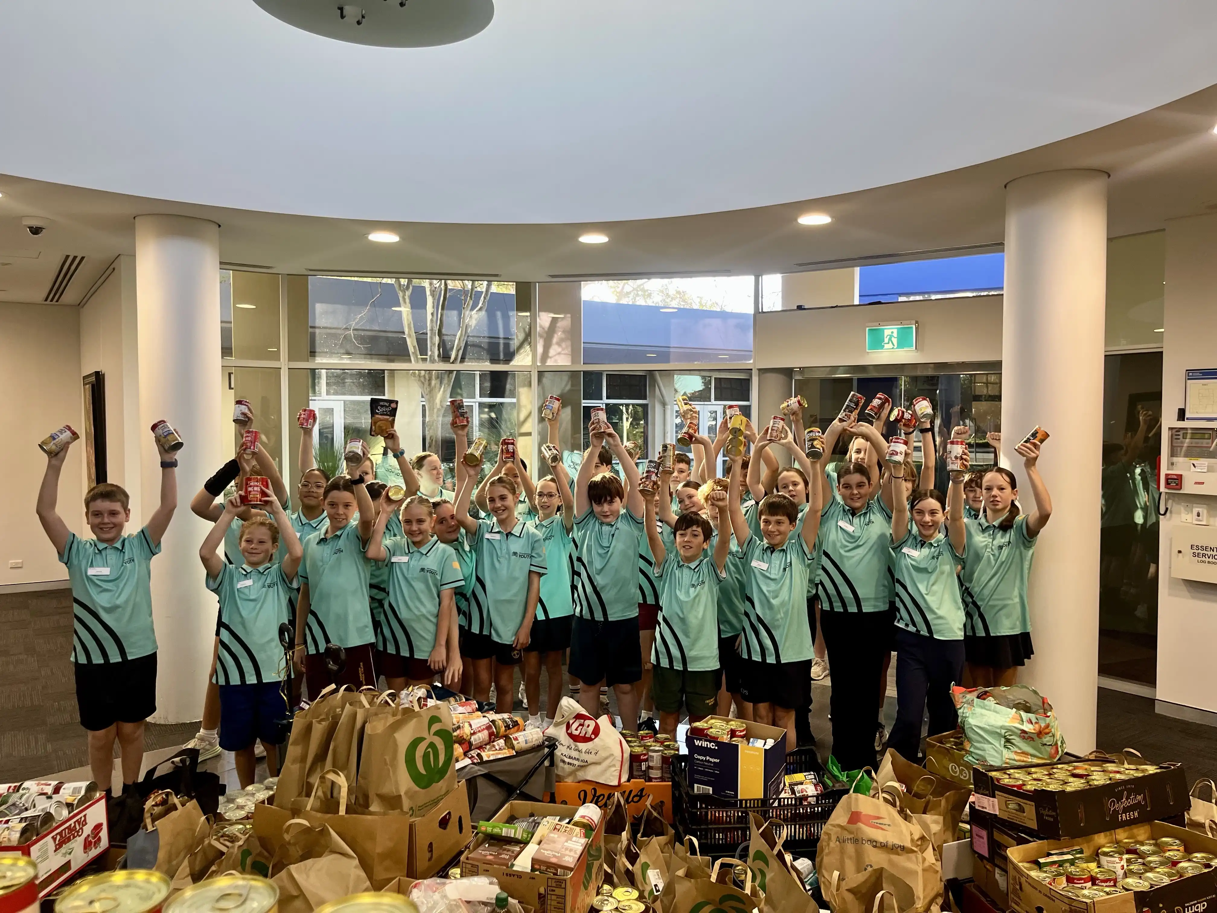 School students hold up donated food items in front of a large collection of community food donations.